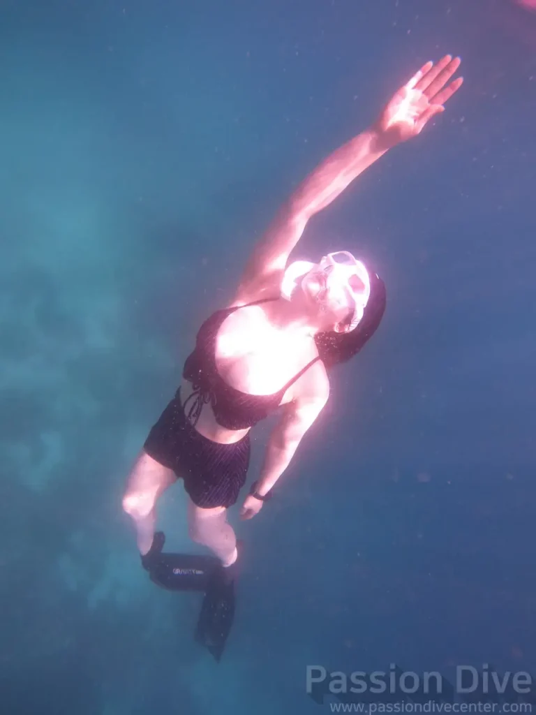 A freediver gliding underwater with one arm extended upward at the Passion Dive housereef in Cebu